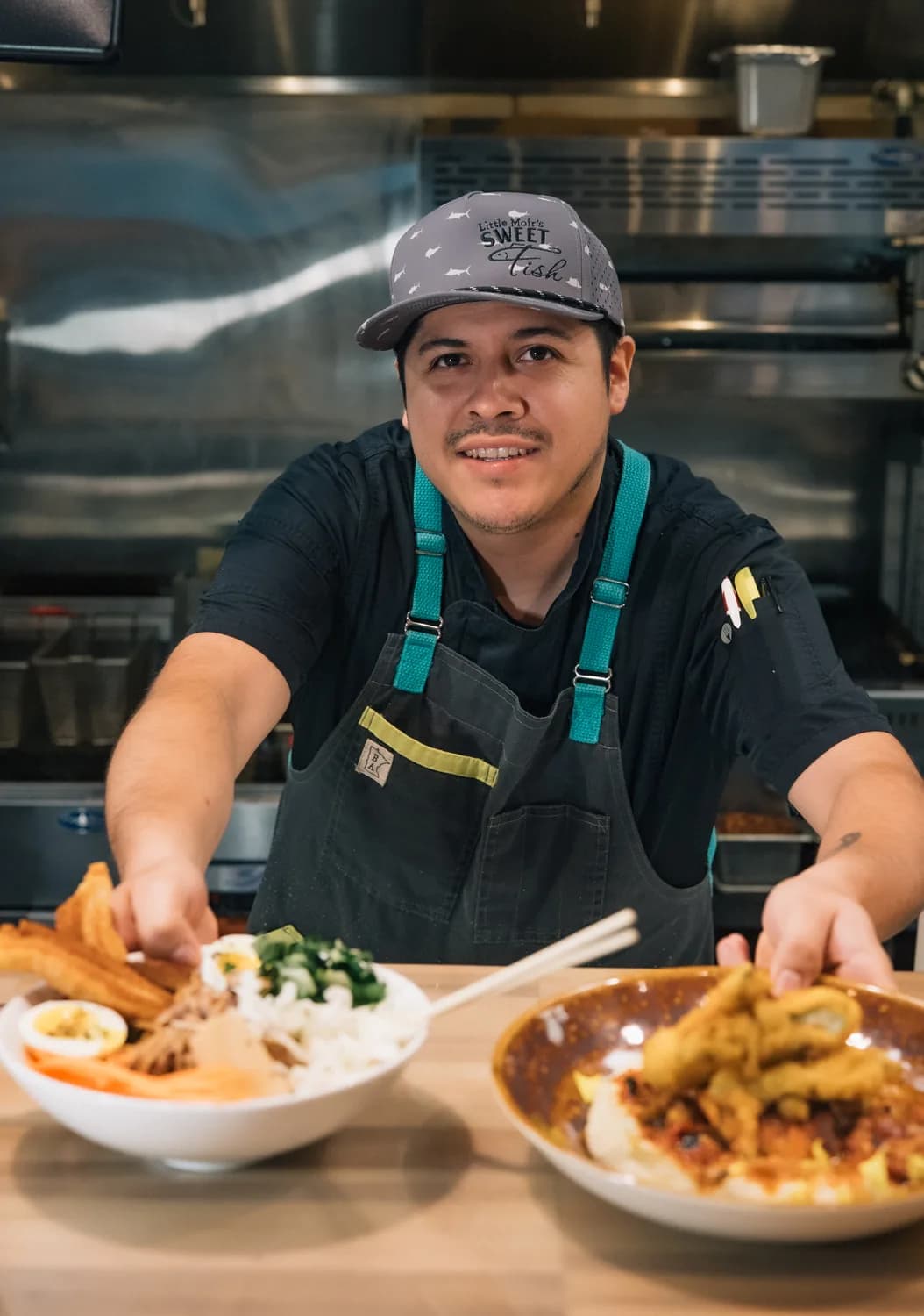 A smiling Sweet Fish chef in a branded cap presenting two bowls at the kitchen pass.