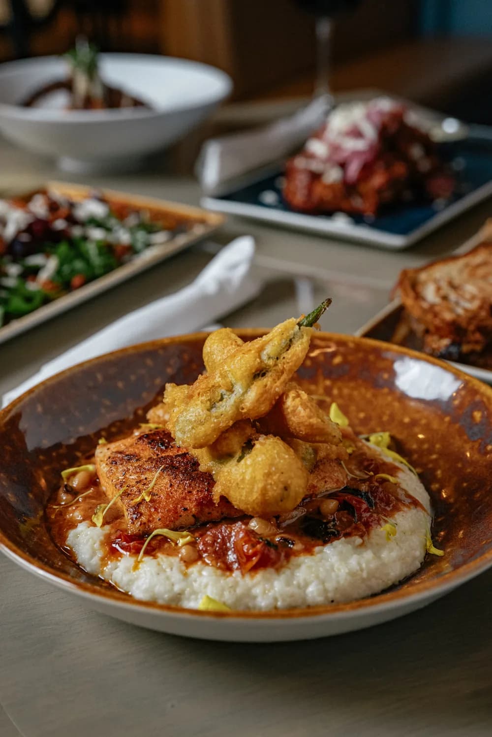 Blackened fish over creamy grits with crispy fried jalapeños and tomato in a speckled bowl.